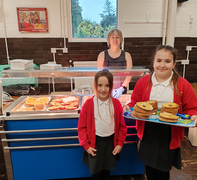 Two Eastover pupils stood in front of the bagel bar in the school hall with Mrs Stevens behind the bagel bar. 